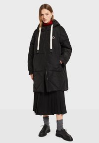 Sort dunjakke med hætte, med hvide snore, diagonalt quiltet mønster og et lille rundt logo på brystet.