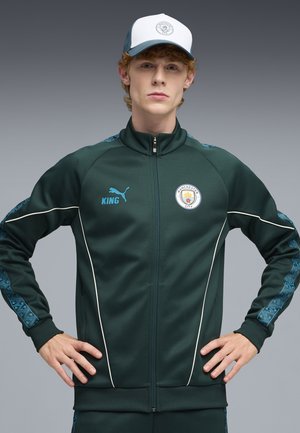 Jeune homme portant une veste de survêtement vert foncé de Manchester City et une casquette assortie, se tenant les mains sur les hanches devant un fond gris.