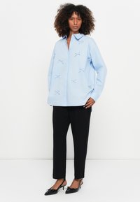 Camisa oversized azul clara com fecho de botão, apresentando laços decorativos. Combinada com calças pretas e sapatos de salto agudo.