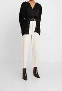 Cardigan nero lavorato a maglia con apertura frontale, abbinato a una blusa nera in satin e pantaloni crema a vita alta. Stivaletti alla caviglia marroni con motivo.