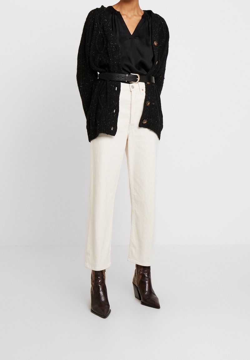 Cardigan nero lavorato a maglia con apertura frontale, abbinato a una blusa nera in satin e pantaloni crema a vita alta. Stivaletti alla caviglia marroni con motivo.