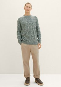 Maglione lavorato a maglia in tonalità blu, colletto rotondo, abbinato a pantaloni cargo beige con tasche e stivali brown con lacci. Texture complessiva liscia.