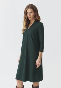 Robe verte à col en V avec des manches trois-quarts, en tissu doux, coupe décontractée et longueur genou, associée à des bottes brunes montantes.