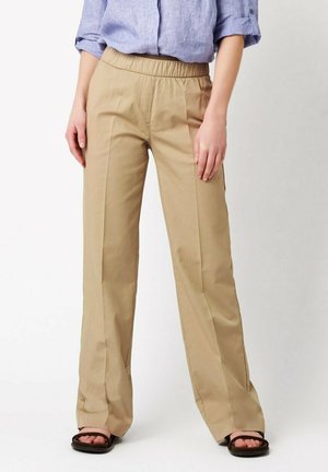 Persona che indossa pantaloni beige a gamba dritta con vita elasticizzata, camicia azzurro chiaro a maniche lunghe arrotolate e sandali neri con punta aperta.