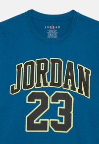 Modra športna majica s kratkimi rokavi z velikim črnim in rumenim napisom "JORDAN" ter številko "23" na sprednji strani.