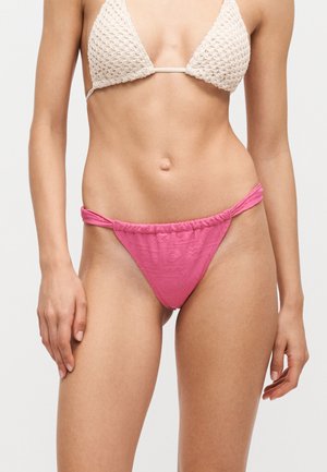 Kobieta ubrana w kremowy szydełkowany top od bikini i różowe majtki bikini z marszczonymi bokami, pokazana od talii do górnej części ud.