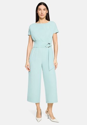 Nő világoskék, rövid ujjú, öves overált visel, amely széles szárú nadrággal párosul, valamint fehér, hegyes orrú magassarkú cipőt.