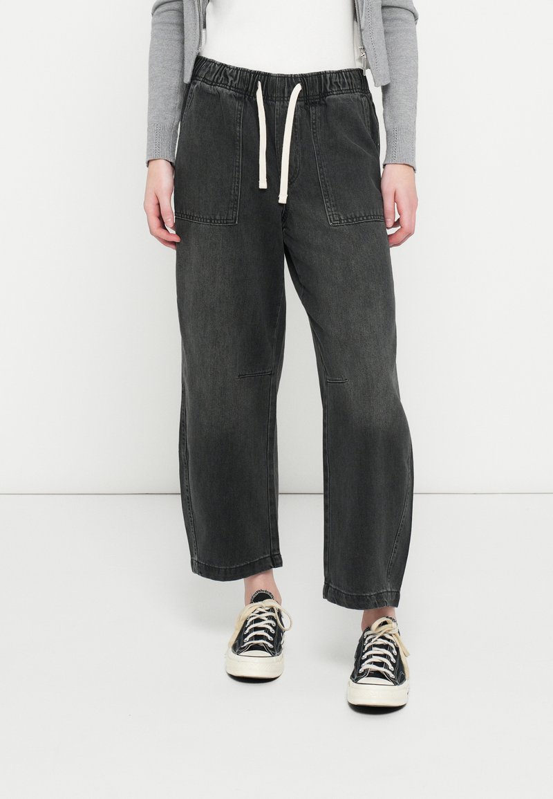 Σκούρο γκρι τζιν παντελόνι jogger με ελαστική μέση και κορδόνι, με δύο μπροστινές τσέπες και φαρδιά γραμμή. Φοριέται με ασπρόμαυρα αθλητικά παπούτσια.