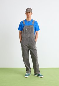 Salopette grise avec bretelles réglables, poches avant, t-shirt bleu et baskets teal. Détails supplémentaires : une casquette grise et un bracelet de perles.
