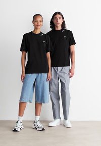 To personer iført svarte Lacoste t-skjorter; den ene i lyseblå denimshorts og sneakers, den andre i stripete bukser og hvite sko, står side ved side.