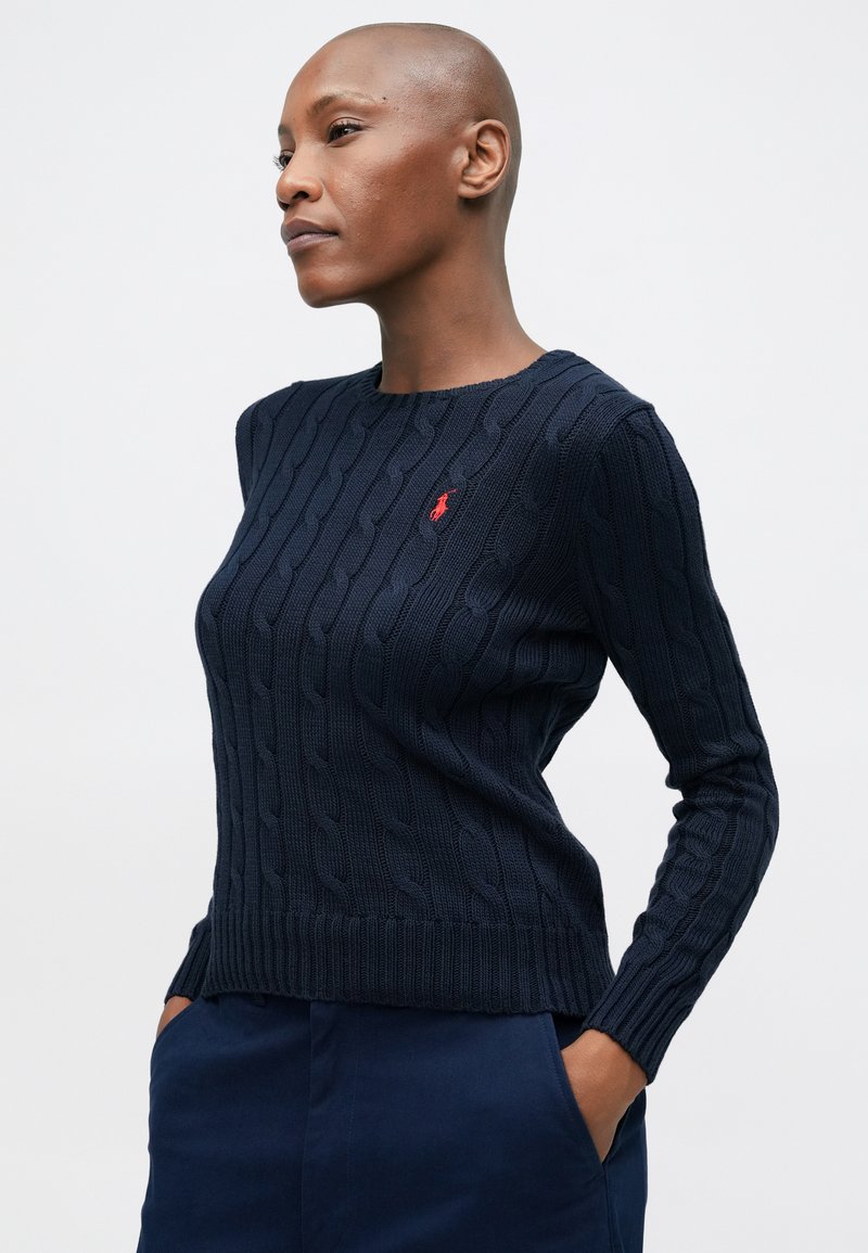 Pull en maille torsadée bleu marine avec un col rond, présentant un motif texturé et un petit logo rouge sur la poitrine gauche. Bords côtes aux poignets et à l'ourlet.