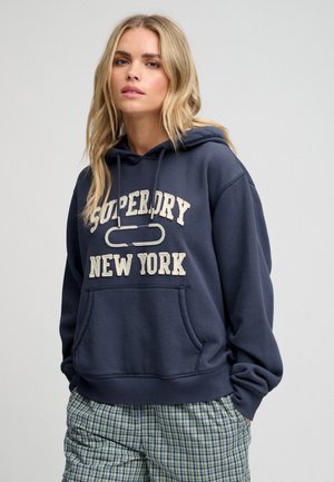 Kobieta z długimi blond włosami, ubrana w granatową bluzę z kapturem Superdry New York i zielone spodnie w kratę na jednolitym jasnym tle.