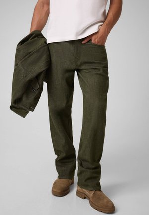 Le modèle porte un jean droit vert olive foncé, un tee-shirt blanc et des bottes marron, et tient un jean assorti devant un fond gris uni.