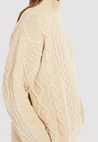 Jersey de punto beige con cuello alto, patrones texturizados y un corte holgado. Los detalles notables incluyen diseños trenzados y en forma de diamante.