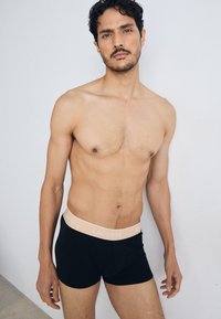Shorts en coton noirs avec une ceinture élastique rose pâle arborant le logo "LACOSTE". Le mannequin a les cheveux courts et foncés et est torse nu.