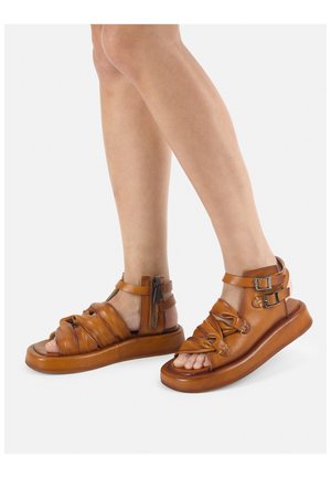Brown leather sandals with knotted design, adjustable ankle straps, and side zipper. Thick sole with a rounded shape. Open-toe style.