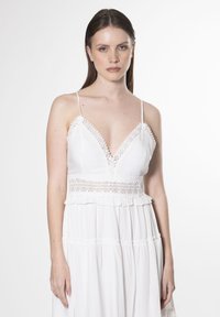 Vestido blanco con borde de encaje, escote en V y tirantes finos; presenta un dobladillo con volantes y una sutil textura en la tela. Detalles de diseño elegantes en todo el vestido.