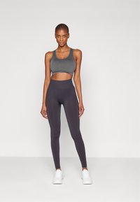Ensemble de brassière de sport grise et de leggings foncés. La brassière présente un design cropped avec des bretelles larges ; les leggings sont taille haute avec une texture lisse.