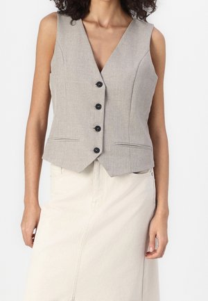 Vrouw draagt een lichtgrijs mouwloos gilet met knopen, gecombineerd met een hoog getailleerde crèmekleurige rok, staand tegen een effen witte achtergrond.