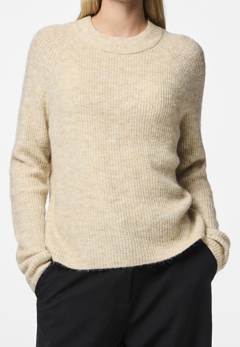 Beige stickad tröja med ribbad struktur, buren med svarta byxor, händerna avslappnat nedlagda i fickorna, visad från halsen till höfterna.