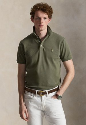 Young man with curly hair wearing olive green polo, white pants, brown belt, and wristwatch, standing against a plain beige background.