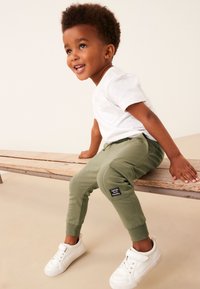 Zelene trenirke s elastičnim manžetnama i zakrpnim džepom, uparene s bijelom majicom i bijelim tenisicama, prikazane na drvenoj klupi.