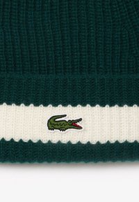 Berretto lavorato a maglia di un verde intenso con una fascia a coste color crema. Presenta un piccolo logo ricamato di coccodrillo con accenti verdi e rossi.