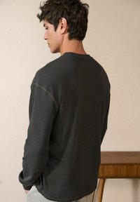 Zwarte ribbelige longsleeve shirt met een ronde halslijn, voorzien van contrasterende stiksels en een ontspannen pasvorm. Draagt door een persoon, gezien van achteren.