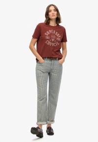 Camiseta de manga corta color marrón con un gráfico circular; combinada con pantalones de rayas de corte holgado y mocasines oscuros.