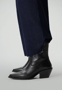 Bottes en cuir noir à mi-mollet avec des bouts pointus et des talons carrés, portées sous un jean large bleu foncé avec des ourlets effilochés.