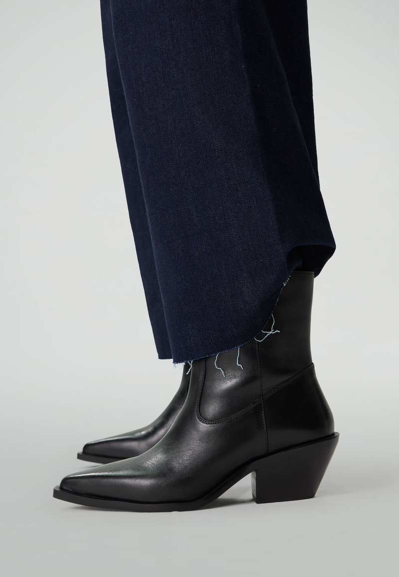 Bottes en cuir noir à mi-mollet avec des bouts pointus et des talons carrés, portées sous un jean large bleu foncé avec des ourlets effilochés.