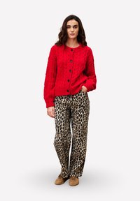 Une femme porte un cardigan en tricot torsadé rouge vif avec des boutons, un pantalon large à imprimé léopard et des chaussures beiges, se tenant debout avec une main dans la poche.