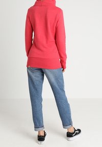Sudadera roja con capucha y un corte relajado, fabricada con un tejido suave. Combinada con jeans claros de tiro recto y zapatillas negras.