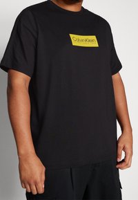 Černé bavlněné tričko s žlutým obdélníkovým logem, na kterém je na přední straně napsáno "Calvin Klein Established 1968". Krátké rukávy, kulatý výstřih.