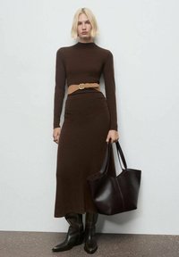 Abito lungo marrone a maniche lunghe con vita aderente e orlo a campana, abbinato a una cintura color sabbia e una borsa a mano marrone scuro, indossato con stivali neri al ginocchio.