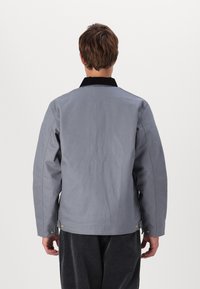 Veste grise avec un col en velours côtelé noir, des poignets boutonnés et une texture lisse. Design simple avec une finition soignée.