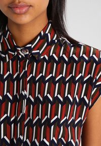 Chemise à manches courtes avec un motif géométrique rouge, blanc et marine sur un fond vert, dotée d'un col classique et d'une fermeture à boutons.