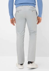 Man in een lichtgrijze broek, witte sneakers met zwart hieldetail, en een blauwe trui, van achteren gezien tegen een witte achtergrond.