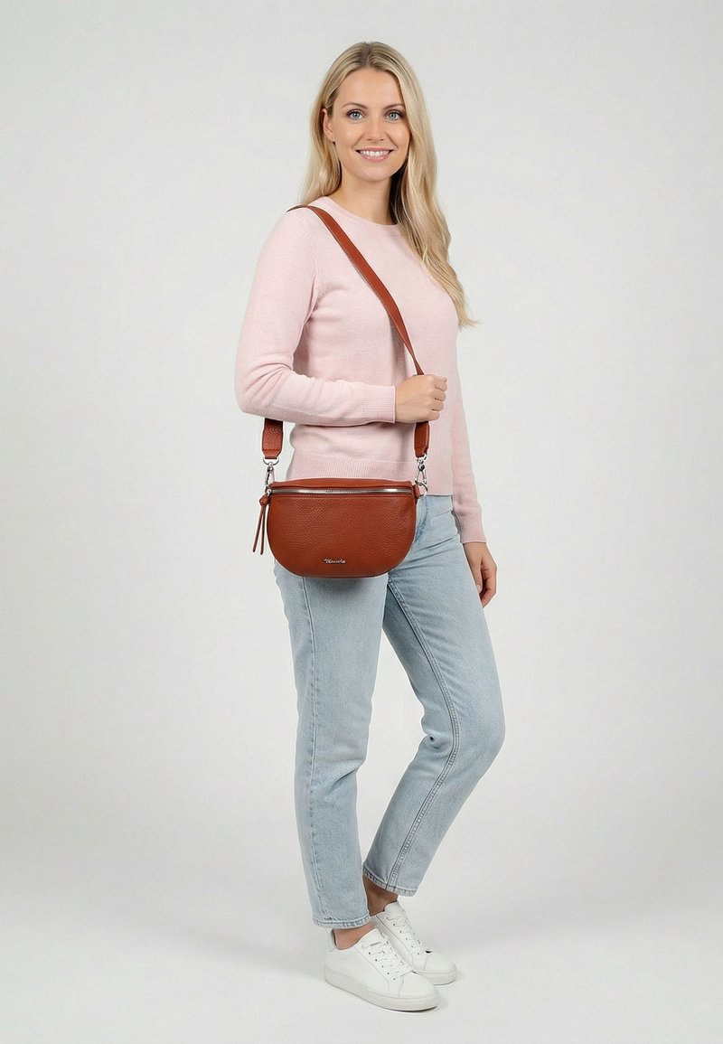 Frau mit langen blonden Haaren, die einen rosa Pullover und helle Jeans trägt, eine braune Leder-Umhängetasche und weiße Sneakers bei sich hat, lächelnd.