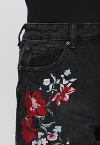 Černé džíny z denimu s živou květinovou výšivkou v červené, růžové, bílé a zelené barvě, hladkou texturou a tradičním designem s pěti kapsami.