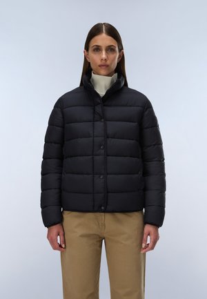 Woman wearing a black padded jacket over a white turtleneck sweater and beige pants, standing against a plain light background.