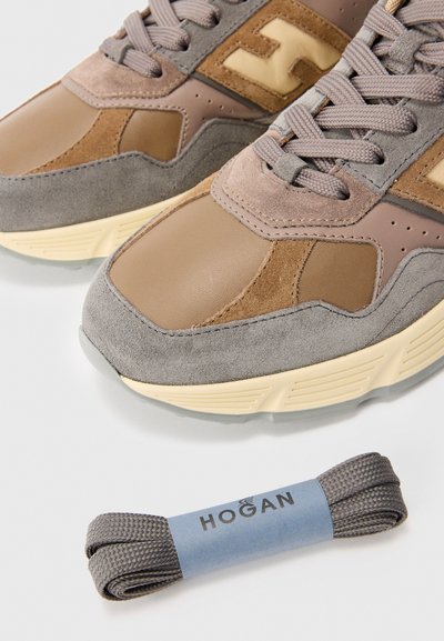 Close-up of brown and gray suede sneakers with beige soles and gray shoelaces beside extra gray laces labeled "Hogan" on white surface.