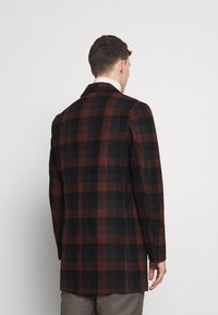 Kabát z vlněné směsi s černo-hnědým kostkovaným vzorem, klasickými klopami, rovnými liniemi a bočním rozparkem, nošený s kostkovanými kalhotami.