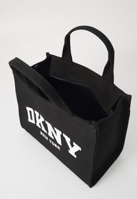 Sac fourre-tout noir en toile avec une forme rectangulaire, doté d'une fermeture zippée et de deux poignées supérieures. L'avant arbore un grand texte blanc "DKNY NEW YORK".