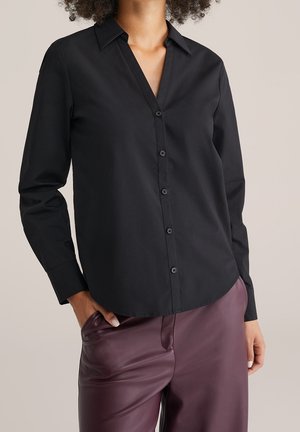 Femme portant une chemise noire à manches longues boutonnée, associée à un pantalon en cuir violet foncé taille haute, main dans la poche.