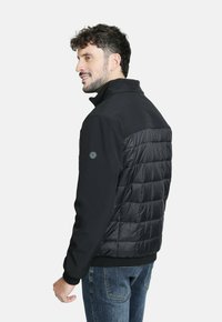 Chaqueta negra con espalda acolchada y con relleno, frente negro sólido, cuello alto y tela texturizada. Presenta un logo en la manga y puños acanalados.