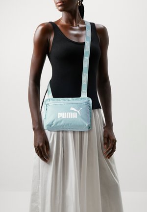 Femme portant un débardeur noir et une jupe blanche, avec un sac bandoulière Puma bleu clair à bandoulière marquée traversant sa poitrine.