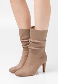 Botas de tobillo beige de ante suave, con punta redondeada y tacón afilado de 7 cm. La parte superior tiene una textura gathered cerca de la abertura.