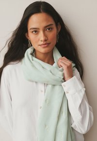 Mujer con cabello largo y oscuro que lleva una camisa blanca y una bufanda verde claro con motivos de hojas doradas, sosteniendo la bufanda cerca del hombro, pendientes dorados.