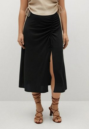 Sort midilengde nederdel med sidesamlinger og en frontslids. Kombineret med brune remsko med hæl og en beige top. Glat stofstruktur.